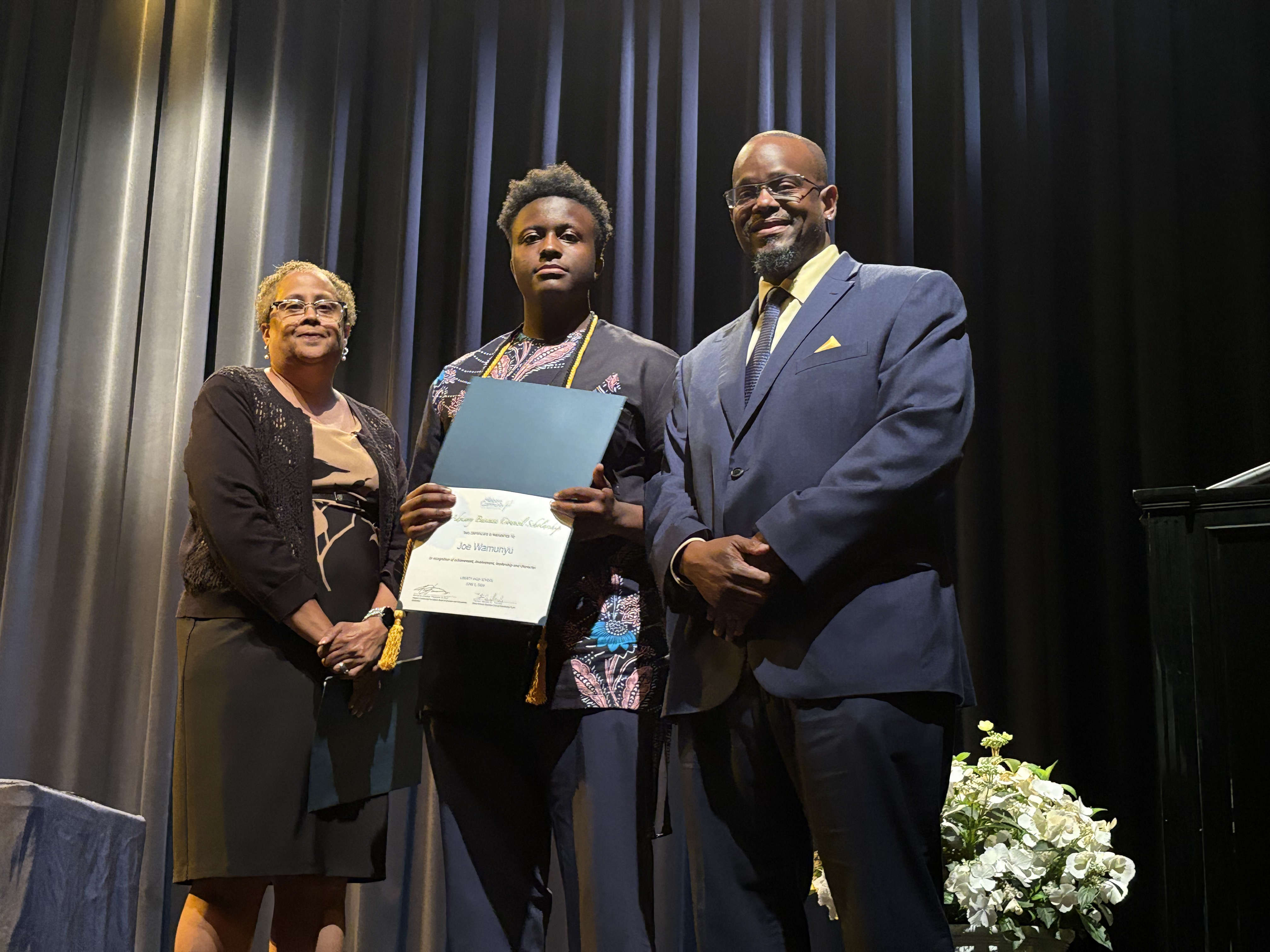 Joe Wamunyu, 2026 BABC Scholarship recipient from Liberty High School smiling proudly on stage with his certificate alongside presenters.